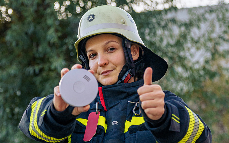 Eine Feuerwehrfrau zeigt den Rauchmelder "Daumen hoch".
